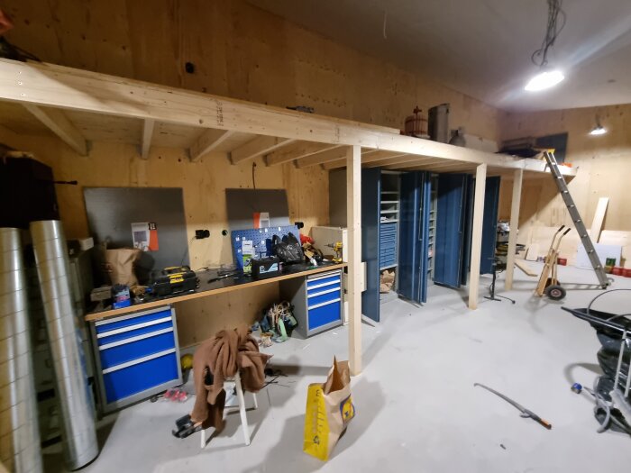 Verkstad med arbetsbänk, verktyg och material; hylla ovan för förvaring. Stökig miljö med pågående renovering och en stege för tillträde.