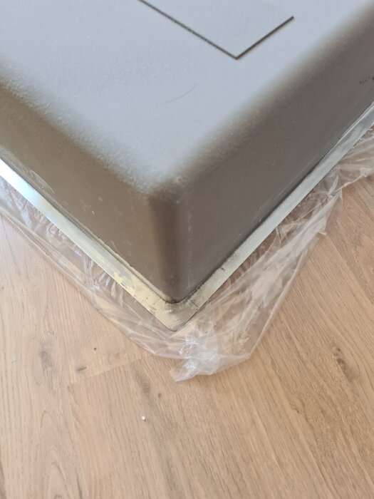Grått föremål limmat med silikon, stående på en plasttäckning över trägolv.