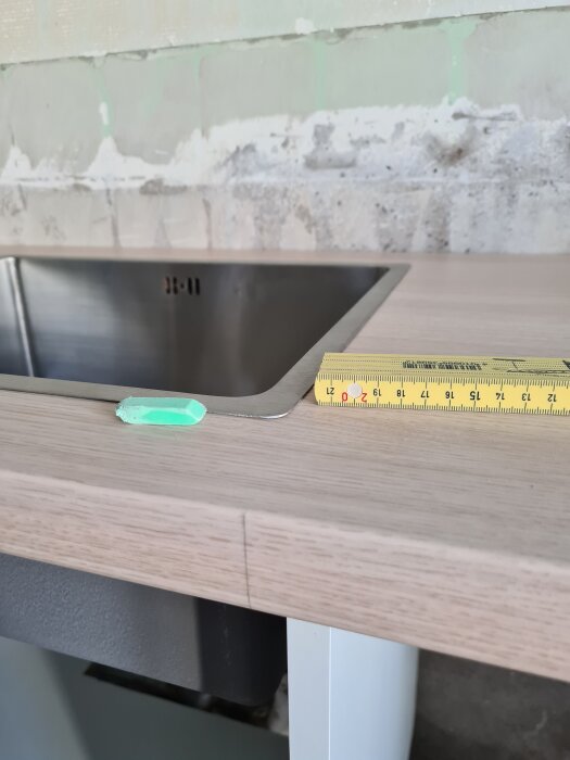 Närbild av en diskbänk med träbänkskiva, ett måttband placerat på kanten och en liten träkil bredvid vid en byggarbetsplats.