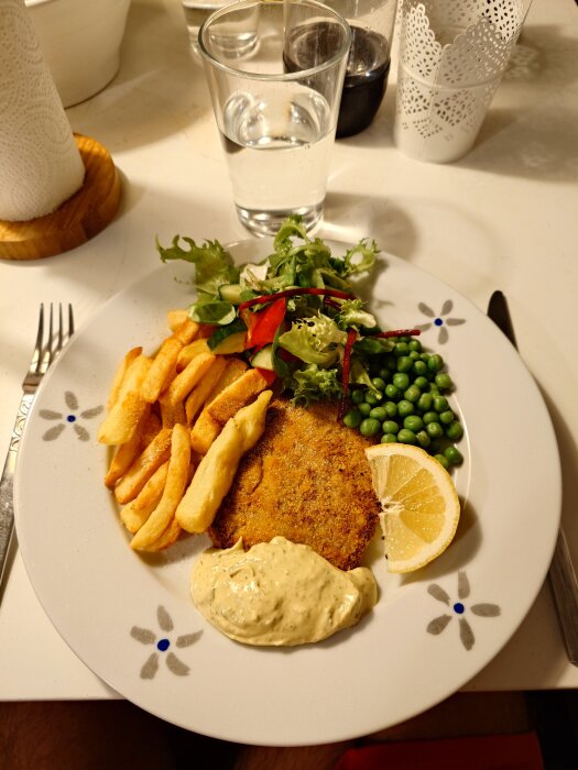 Tallrik med schnitzel, pommes frites, ärtor, citronskiva, béarnaisesås och sallad, serverad på ett vitt bord.
