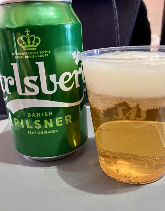 Grön burk med dansk pilsner och ett plastglas med öl, stående på ett bord.