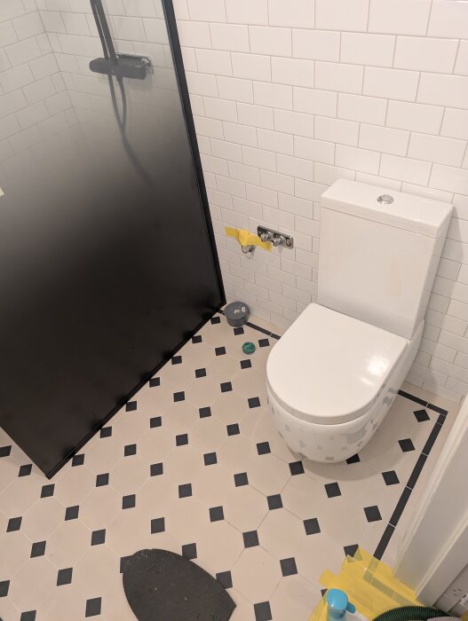 Toalett installerad i ett badrum med vita kakelväggar och svartvitt golv; en dusch i hörnet.