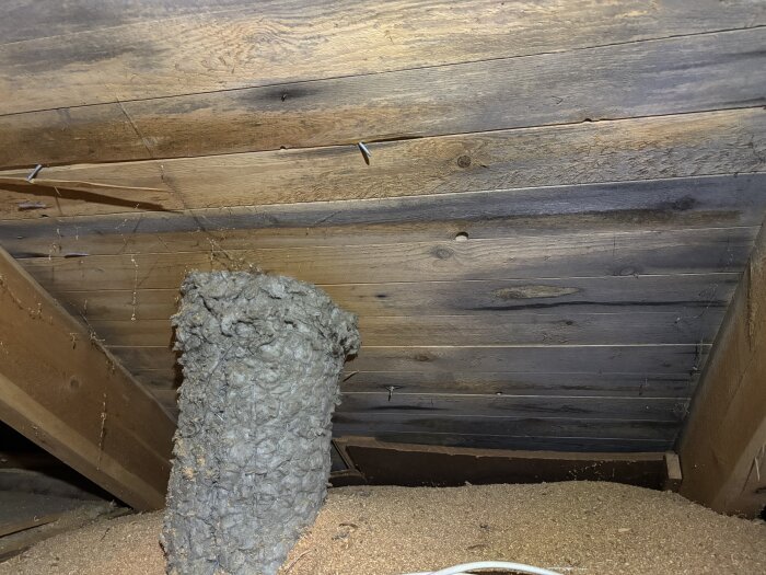 En bild av råspont på en vind, med mörkare partier och en stor ventilationskanal från badrummet, samt synlig sågspån på golvet.