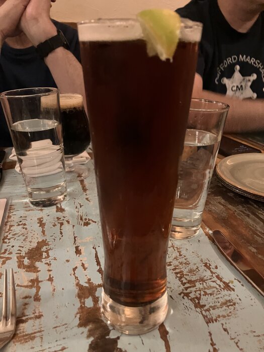 Högt glas med mörk dryck och limeklyfta på kanten, stående på ett nött träbord, omgiven av vatten- och ölglas samt bestick och tallrik.