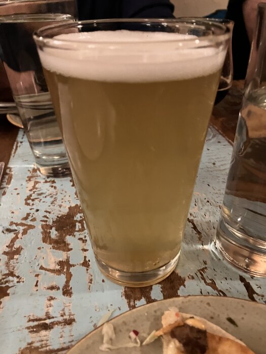 Ett glas ljus öl på ett slitet träbord, omgiven av andra glas och en tallrik med matrester.