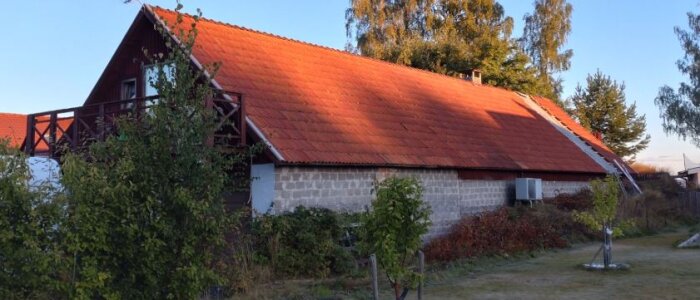Lada med rött tegeltak, omgiven av gröna buskar och träd, i kvällssol. Potentiellt projekt för renovering och isolering för verkstad och gym.