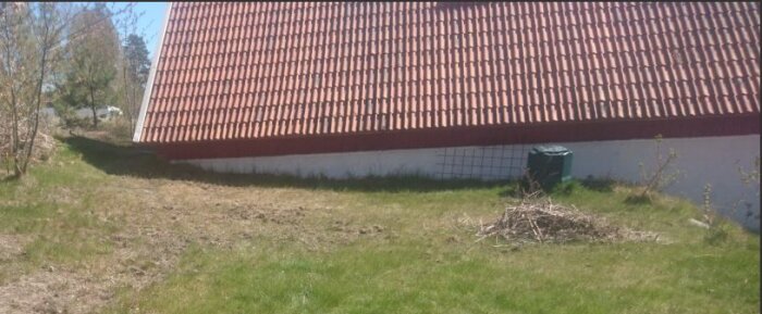 En lada med tegeltak står på en gräsbevuxen lutning. Några buskar syns i bakgrunden samt en hög med kvistar på marken.