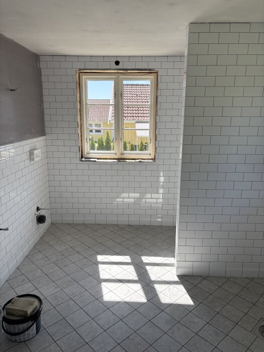 Nyrenoverat badrum med vita kakelplattor på väggarna och grått klinkergolv, sett genom ett fönster med ljusinsläpp.