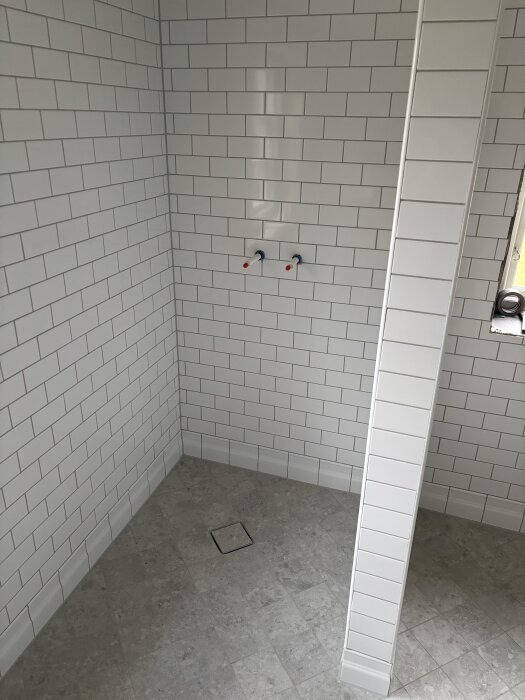 Nytt kaklat hörn av ett badrum med vita plattor och ett grått klinkergolv, samt två vattenuttag på väggen.