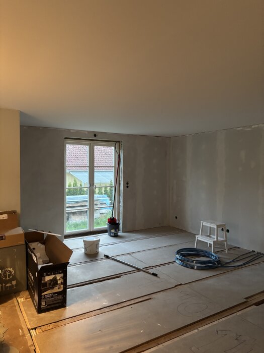 Renoverat rum med nytt tak, verktyg och material på golvet, inklusive en vit stege, kabelrulle och en kartong. Fönster visar trädgård utanför.