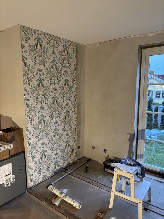 Vägg med blommig tapet bredvid grå vägg, fönster till höger, golvverktyg och stege i rum under renovering.