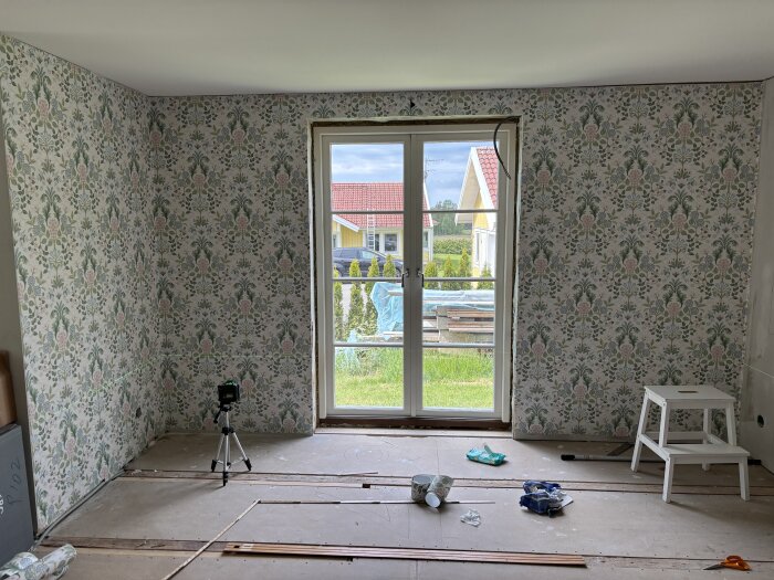 Rum under renovering med nyblommig tapet, dörr till trädgård, verktyg och material på golvet, vit pall nära dörren, och utomhus utsikt.