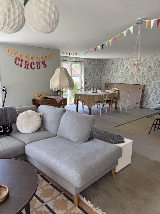 Renoverat rum med grå soffa, matbord, unik taklampa och färgglada vimplar. Väggarna är tapetserade med blommönster.