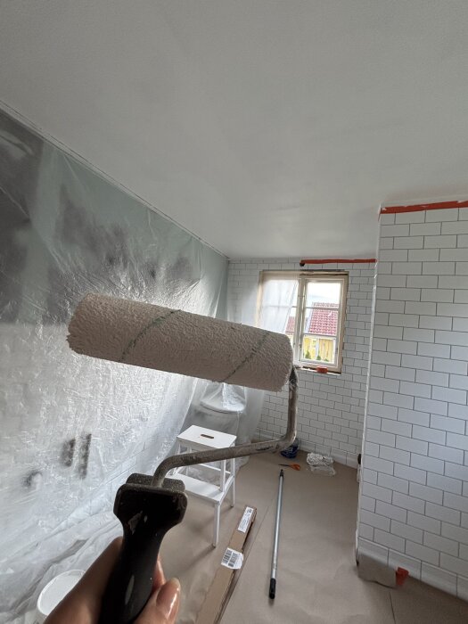 Hand håller en målarroller, omgiven av ett rum under renovering med vita kaklade väggar och täckt golv, ett fönster i bakgrunden.