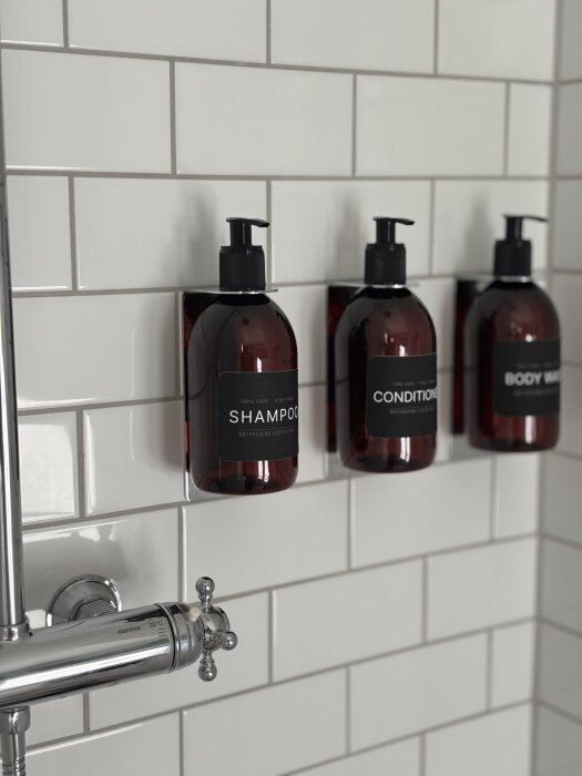 Tre bruna flaskor för schampo, balsam och duschtvål monterade på en vit, kaklad badrumsvägg. En del av en duschkran syns bredvid.