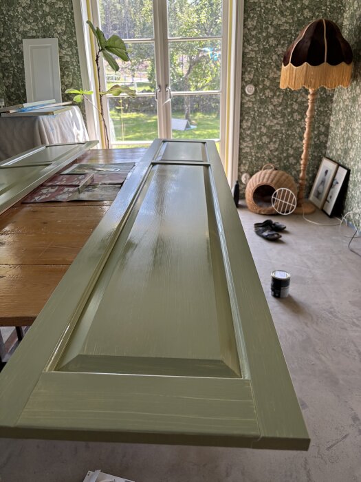 Grönmålad dörrpanel på ett bord framför ett fönster, omgiven av blommönstrad tapet, växt och retro lampa i ett rum.