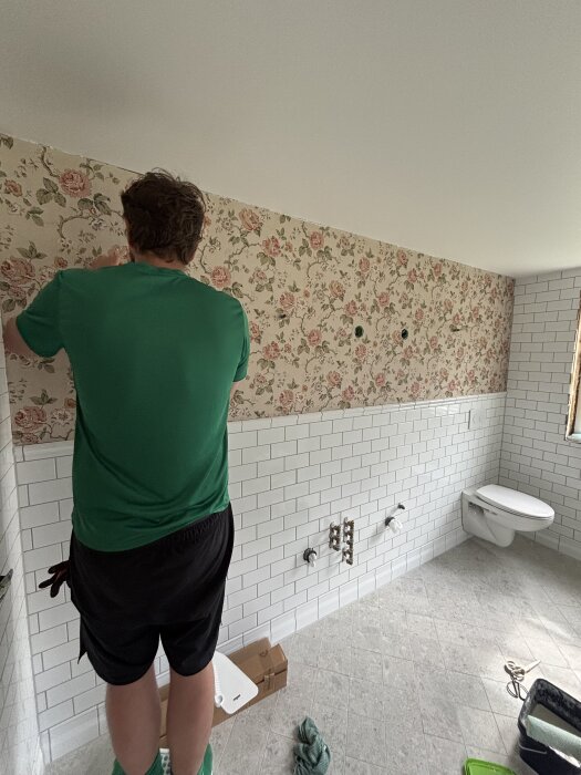 Person tapetserar en vägg i ett badrum med blommigt mönster, över vita kakelplattor. Golvet är flytspacklat och toaletten saknar handfat.