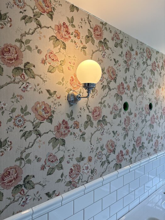 Blommig tapet och kaklad vägg i badrum med en vägglampa ovanför kaklet, del av renoveringsprojekt.