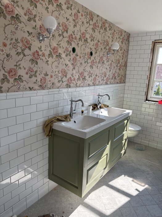 Badrum med blommig tapet, vita kakelplattor på väggarna, grön dubbel handfat med skåp, och ett fönster som släpper in solljus.