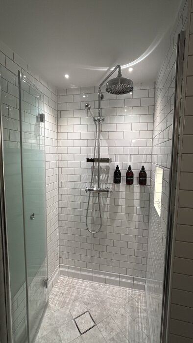 Modernt badrum med vita kakelväggar och dusch med regnduschhuvud. Tre flaskor för schampo och tvål står på en hylla.