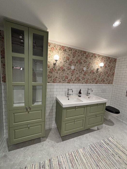 Grönt badrumsskåp, dubbelt handfat med gröna luckor, blommig tapet, vita kakelplattor och randig matta i ett renoverat badrum.