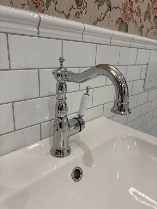 Närbild på en vit handfat med en kromblandare, omgiven av vita kakelplattor och en tapet med blommedesign.