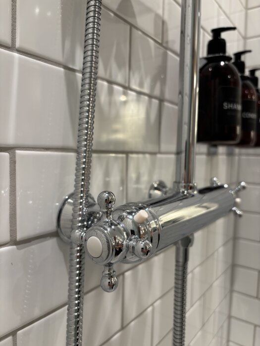 Kromfärgad duschblandare på vit kaklad vägg med schampoflaskor i bakgrunden.