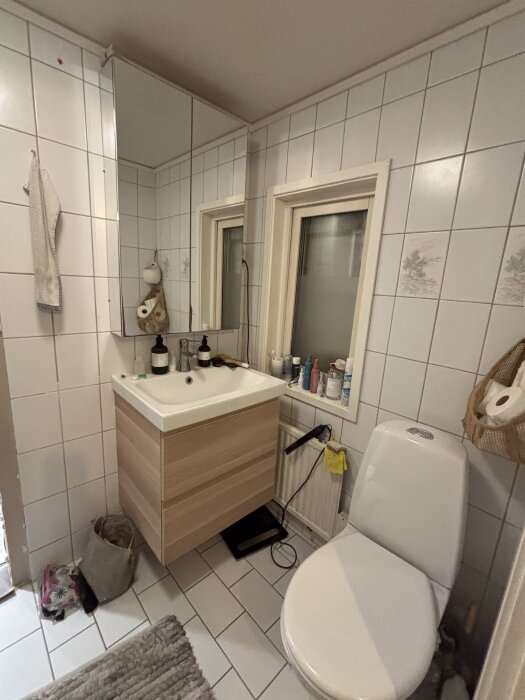 Litet, vitkaklat badrum med spegelskåp och toalett. Vask med träimitation och olika flaskor på hyllan vid fönstret.