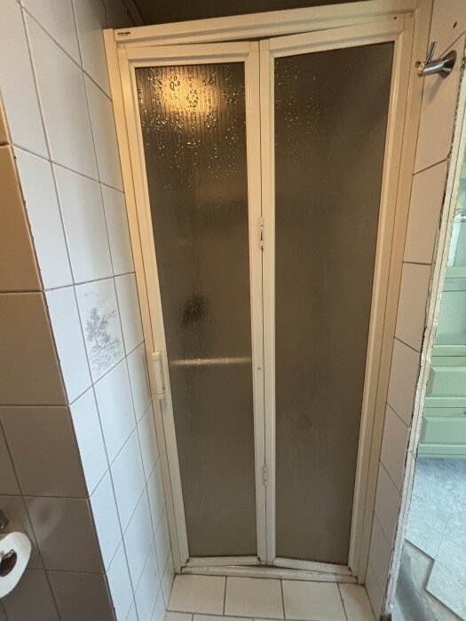 Duschhörna med vita dörrar av plast och frostade glasrutor, omgiven av beige och vita kakelplattor, i ett trångt badrum.