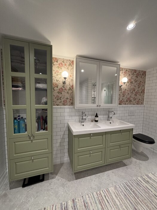 Grönt badrum med dubbla handfat, spegelskåp, blommig tapet och vit kakelvägg. Grön skåpsinredning med förvaring.