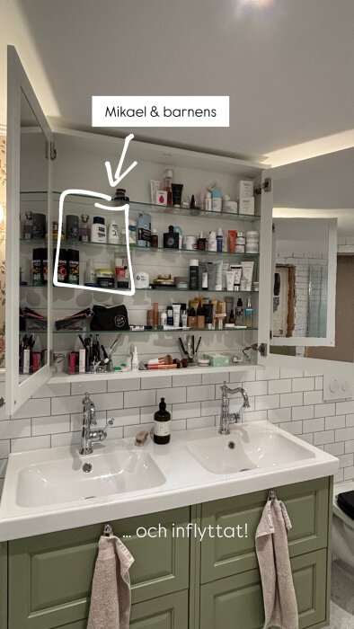 Öppet spegelskåp i badrum med dubbla handfat. Grönt underskåp och hyllor fyllda med hygienprodukter, text "Mikael & barnens" över skåpet.