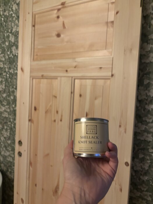 Hand som håller burk med shellack framför en träfärgad innerdörr, del av renoveringsprojekt i badrum.