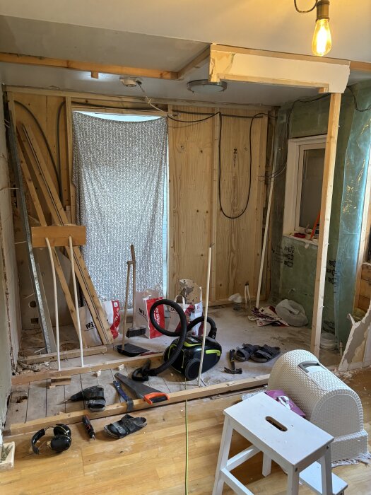 Byggarbetsplats i badrum, verktyg och material på golvet, dammsugare i centrum, ofärdiga väggar och tak, bygglampor och fönster synliga.