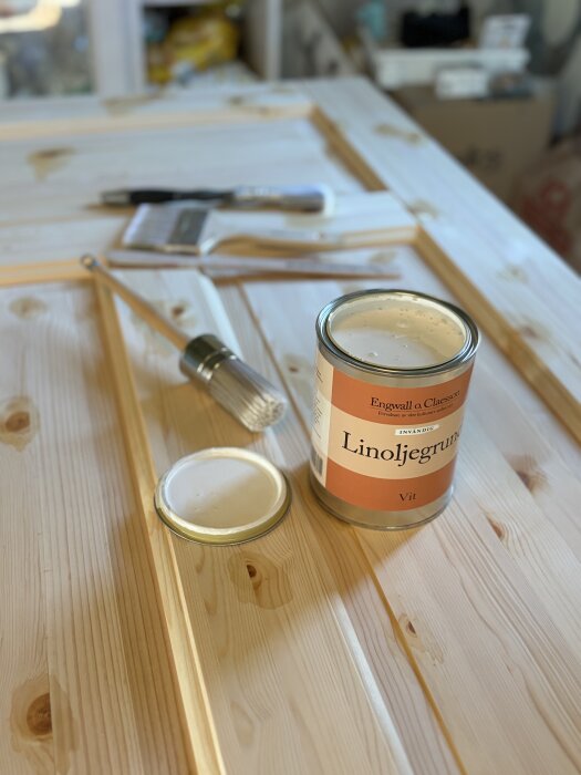 Öppen burk med vit linoljegrundfärg på ett träbord, omgiven av målarpenslar och lock.