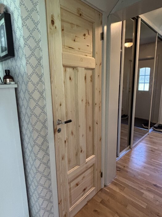 En trädörr i en hall med ljust trägolv och mönstrad tapet. Dörren är av obehandlat trä med metallhandtag. En spegelvägg ses till höger.