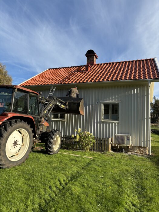 Traktor med skopa vid rött tak på villa, person står i skopan för att nå taket. Klarblå himmel, solbelyst fasad.