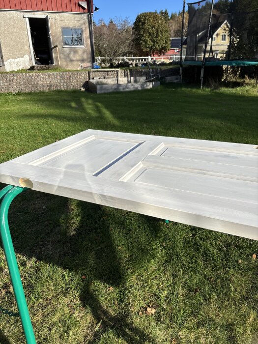 Vitmålad dörr ligger utomhus på ett par färgglada bockar, omgiven av grönska och trädgårdslandskap.