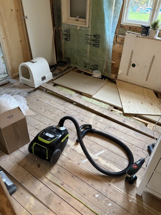 Renoveringsprojekt pågår i badrum med uppbrutet trägolv, isolerade väggar och verktyg som dammsugare.