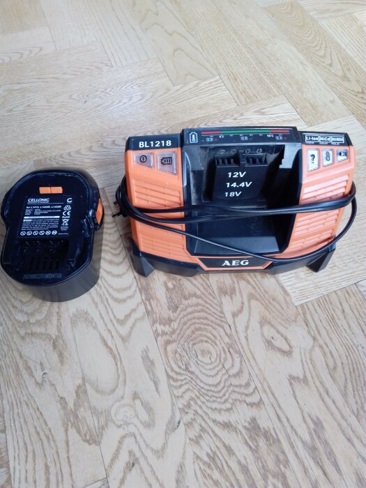 Batteriladdare och batteri av märket AEG på ett trägolv. Laddaren visar inga tecken på fulladdat batteri.