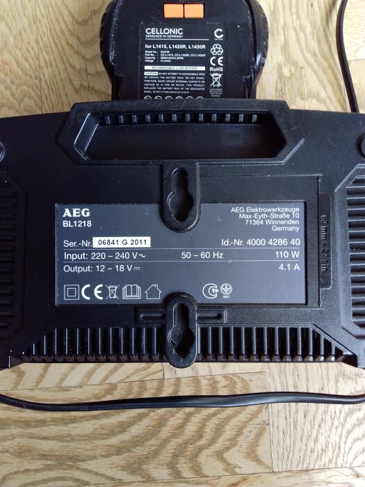 Laddare för AEG BL1218-batteri visas ovanpå trägolv, etiketter med specifikationer synliga. Två batterier av samma fabrikat enligt inlägg.