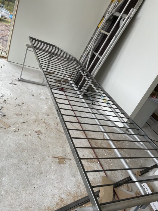 En stor metallram, troligen del av ett byggprojekt, placerad inomhus. Stegar och verktyg syns i bakgrunden. Betonggolv med byggmaterial.
