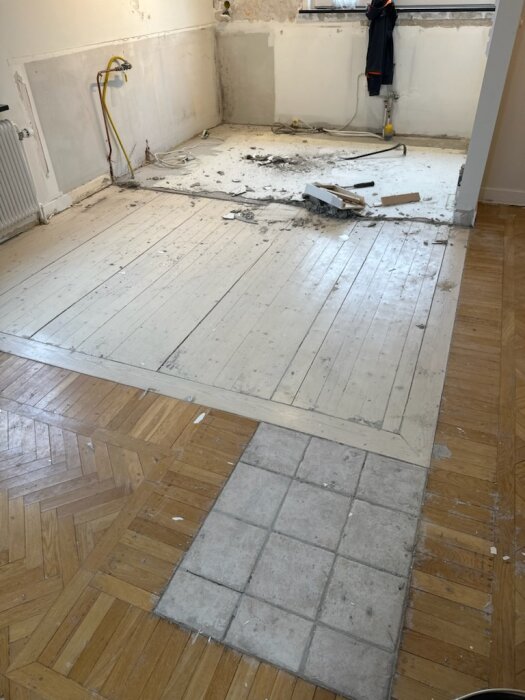 Olika golvmaterial inklusive fiskbensmönstrat trä, vitmålat brädgolv och stengolv i ett rum som ska renoveras.