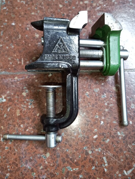 Litet svart och grönt bordsskruvstycke märkt "Made in USSR" på rödbrun stenig yta.