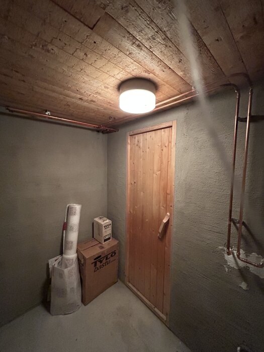 Rum med trädörr, synliga kopparrör i takhöjd och vägg. Förvaring av bastuartiklar. Betongtak och väggar, taklampa lyser.
