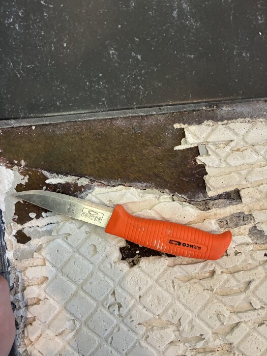 Sprucken grundplatta synlig under borttagna klinkerplattor. En kniv med orange handtag ligger på ytan.