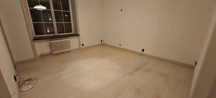 Tomt rum med träplankgolv som visar slitage nära ett fönster med värmeelement; kablar syns på golvet.