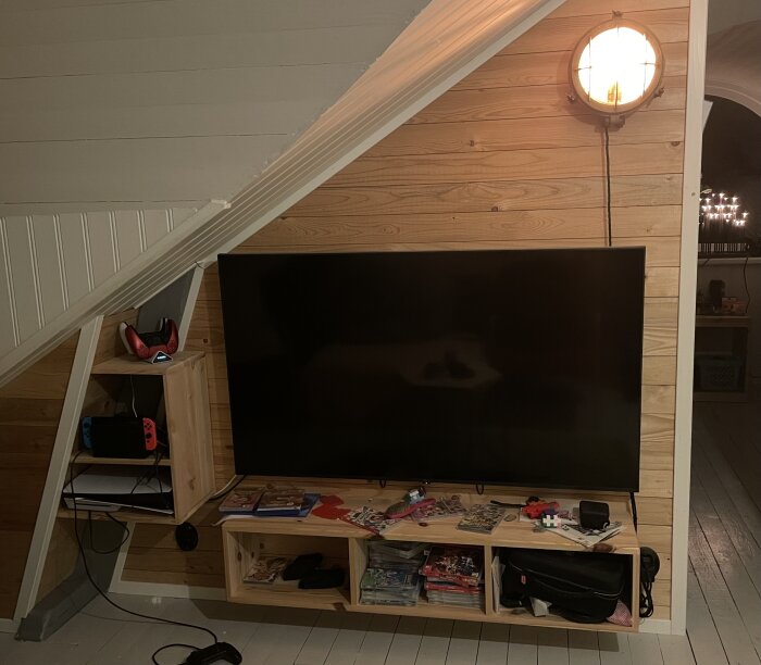 TV-bänk och tv-spelshylla i trä med konsol och spel i barnens tv-rum, belyst av vägglampa.