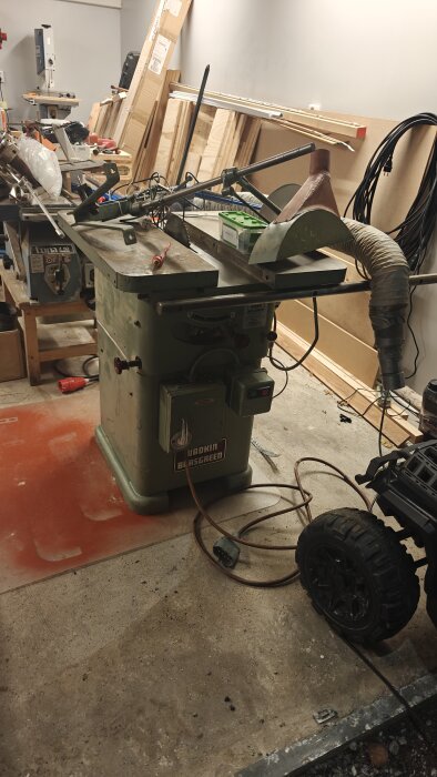 Grön industrisåg med orange kabel och dammsugarslang i garage, omgiven av verktyg och träplankor.