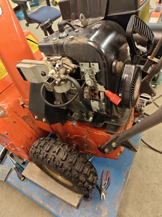 Snöslunga med ett nytt däck och renoverad förgasare, omgiven av verktyg på ett golv i ett garage.