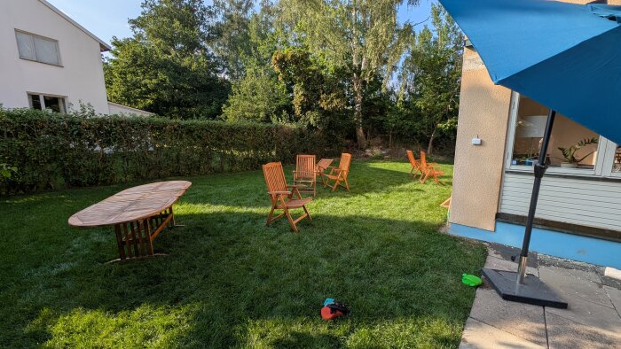 Grön gräsmatta med flera orange utemöbler, ett hopfällt bord och en blå parasoll vid ett hus.